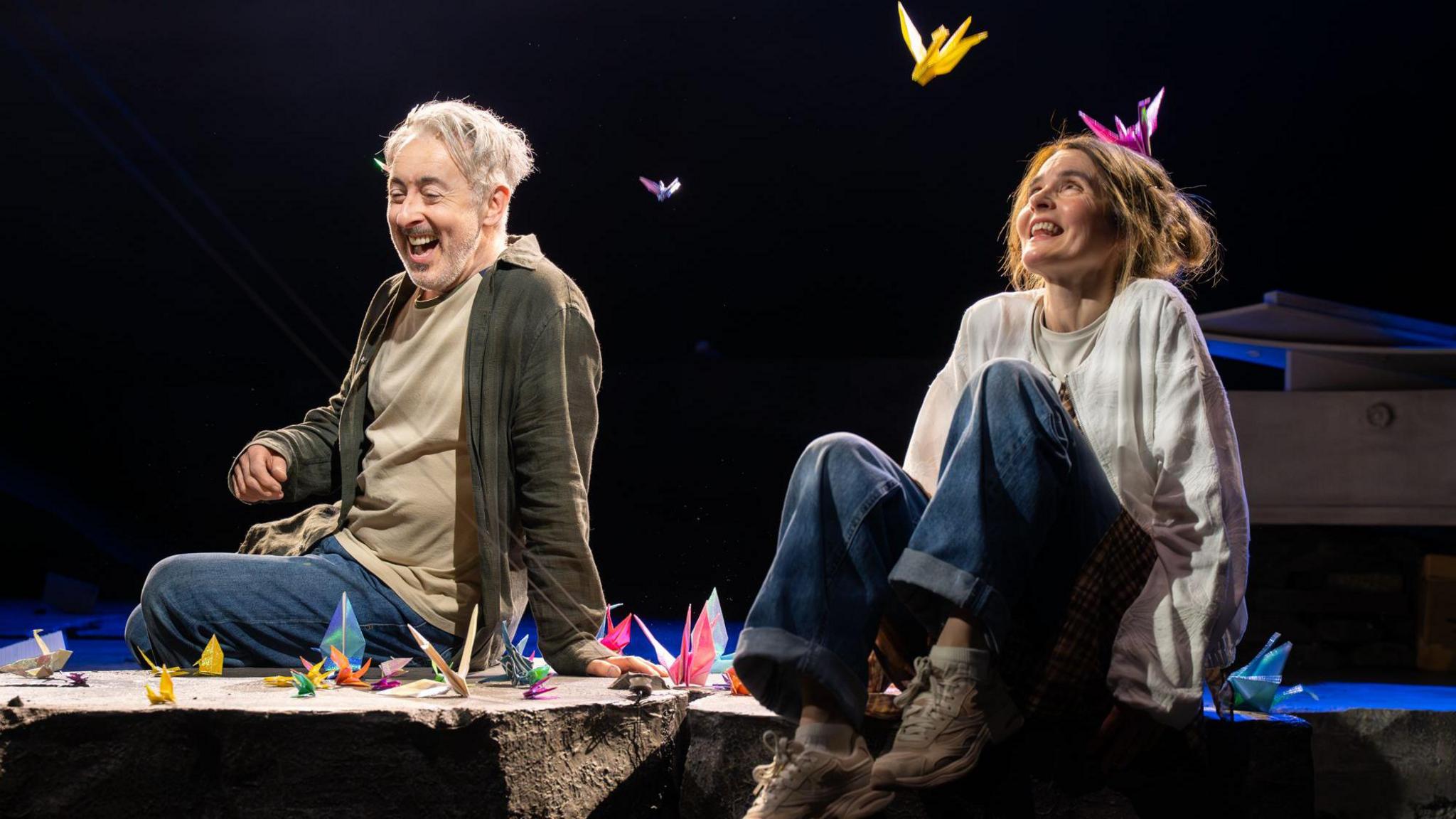Two performers sit on the edge of a stage set, surrounded by brightly coloured origami cranes scattered around and suspended in the air. Dramatic spotlights illuminate the scene against a dark background, drawing attention to the vivid paper sculptures and casual costumes.  The theatrical setting creates a striking contrast between the simple staging and the whimsical, dreamlike display of floating paper birds.