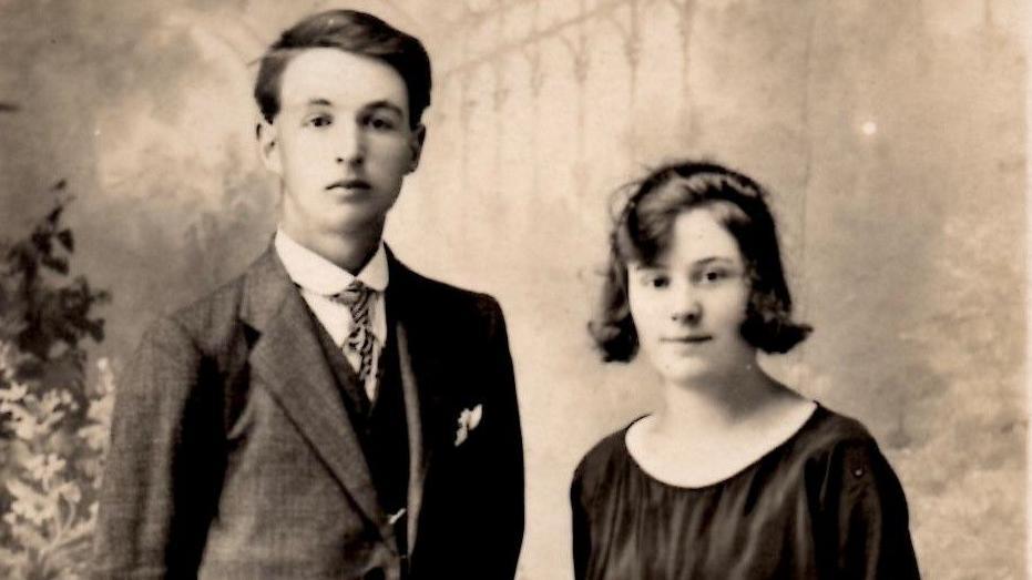 A black and white photograph showing a young man with very dark hair wearing a suit, and a woman with dark hair down to her chin wearing a black blouse.