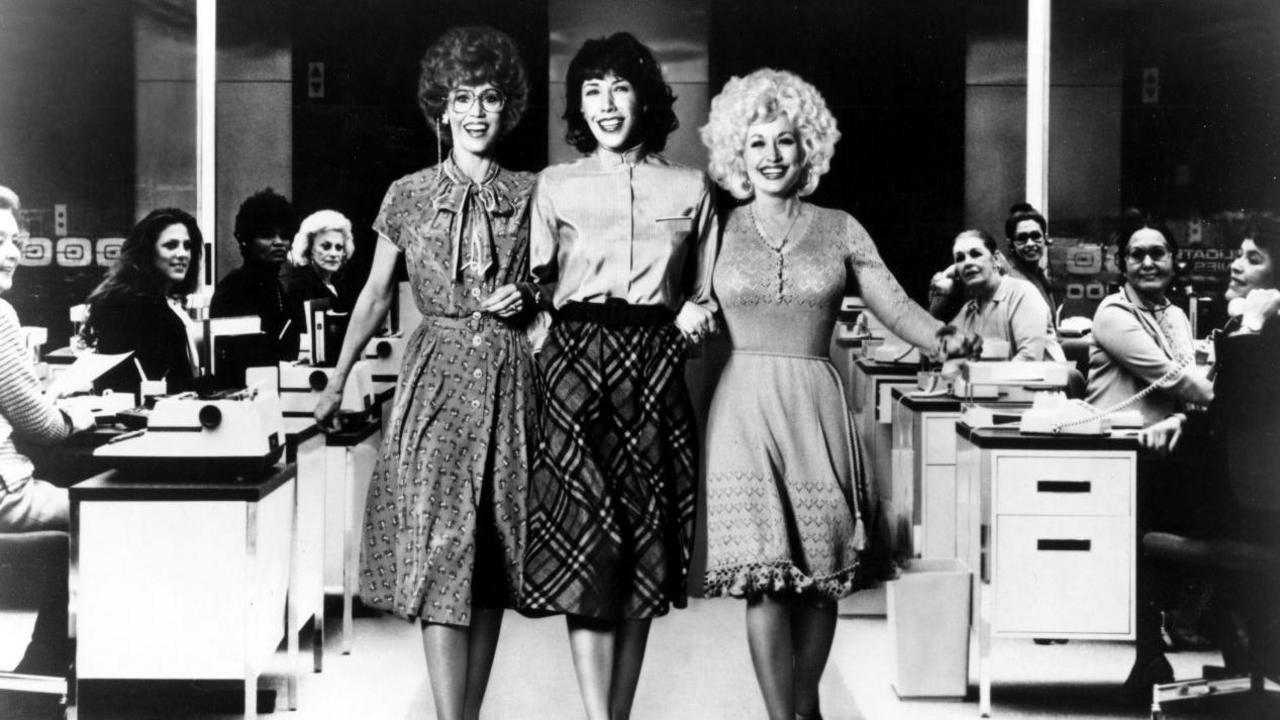 The three link arms and are pictured walking through the office with desks in the background of this black and white photo.  The female typists in the background are all smiling and looking at the three ladies.