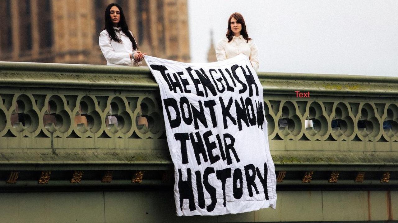 photograph of two women standing on a bridge outside Westminster beside a large banner draped over the railing. The banner reads, “THE ENGLISH DON’T KNOW THEIR HISTORY” in bold, hand-painted capital letters.