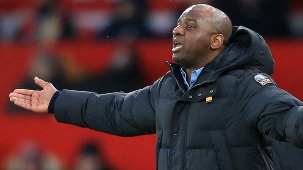 Patrick Vieira, visible from stomach up wearing a thick black padded jacket over a light blue shirt, holds his right arm outstretched at his side as he appeals during a football game. A stand can be seen out of focus behind him in the distance