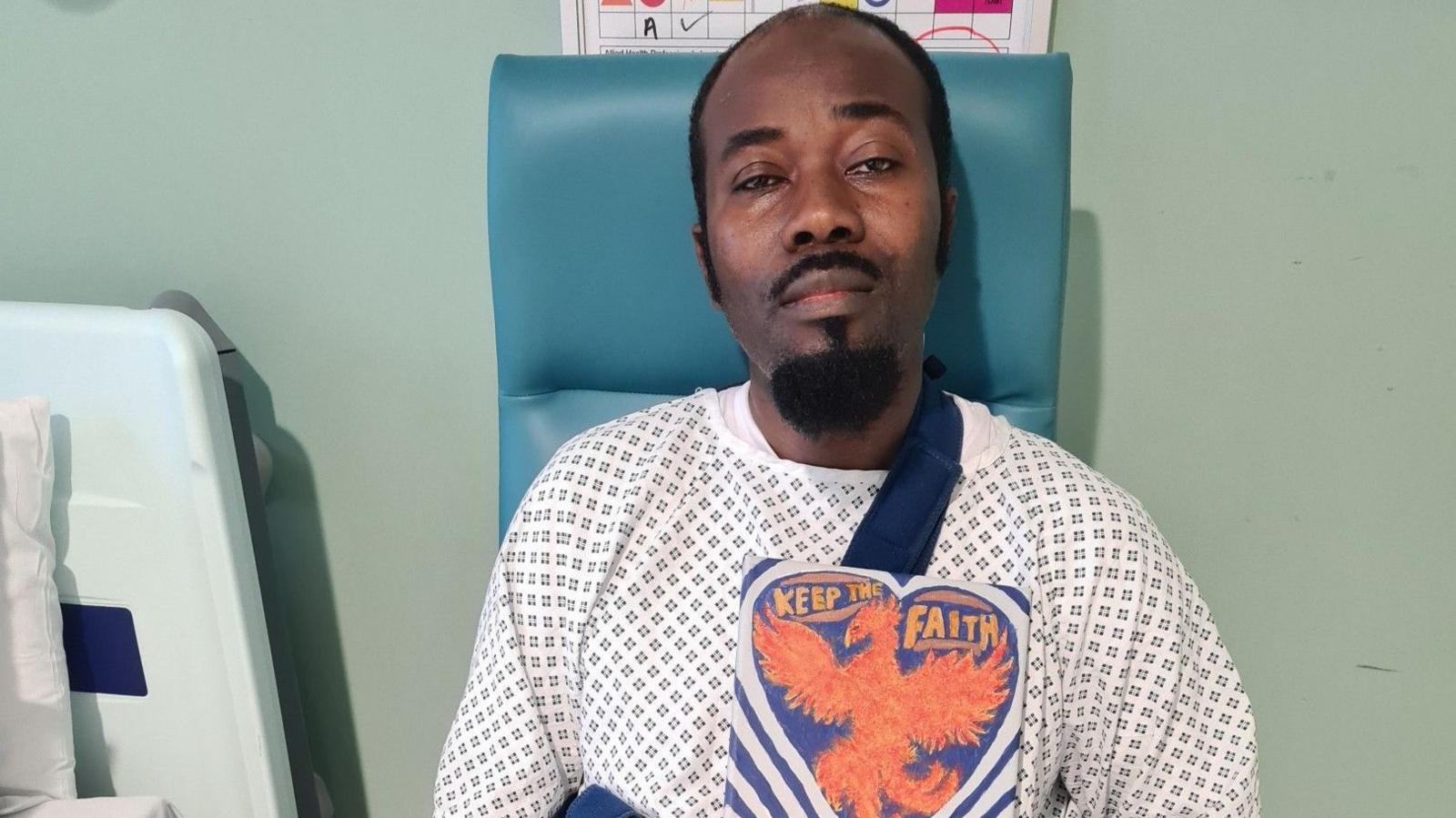 Bernard Agyemang is wearing a patterned hospital gown and is sitting on  a chair. He is holding a painted sign which reads: Keep the faith.