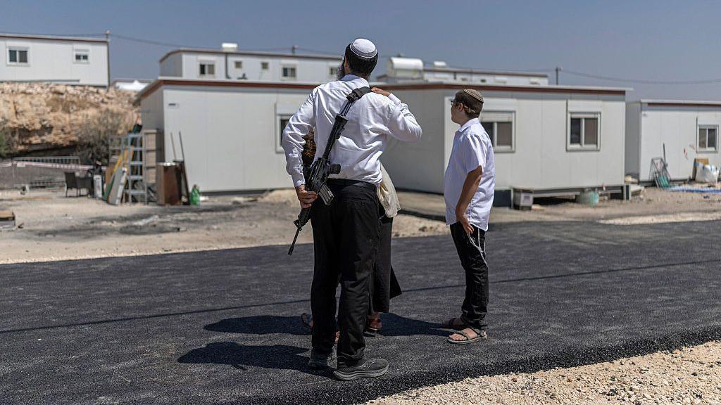 Three Israeli settlers stand outside houses after the reopening of a settlement at Ganim in the Israeli-occupied West Bank on 13 August. One is a man armed with a rifle. The image has a blue border with the BBC Verify logo in the the top left corner.