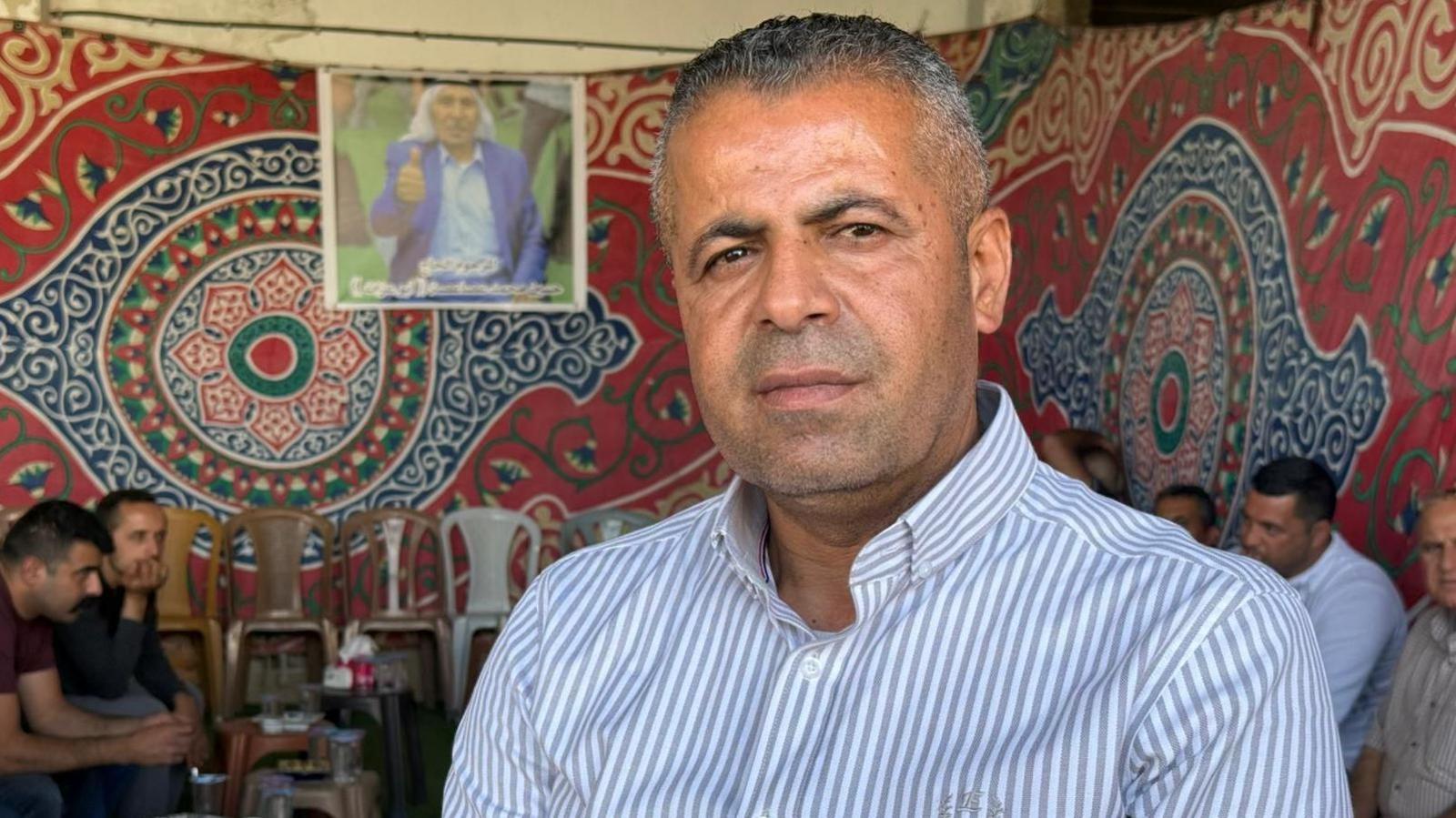 A man in a striped white and blue shirt sits in front of tapestry wall sheets, with people sitting down in the background.