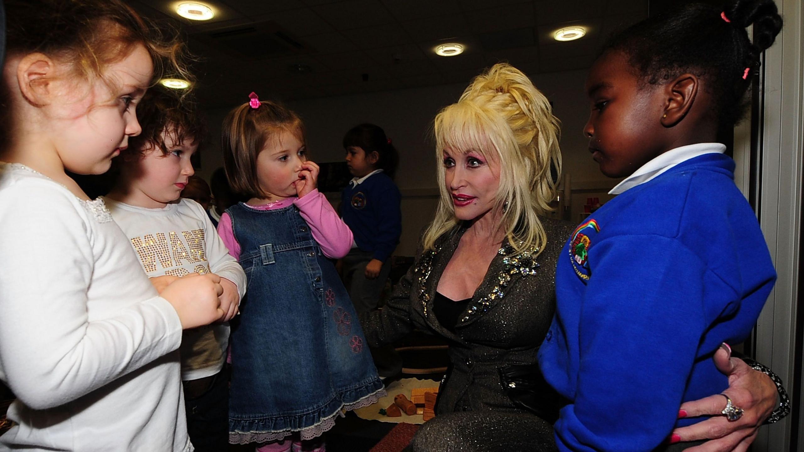 Dolly Parton crouches down and speaks to several children. She has her arms around two of them.