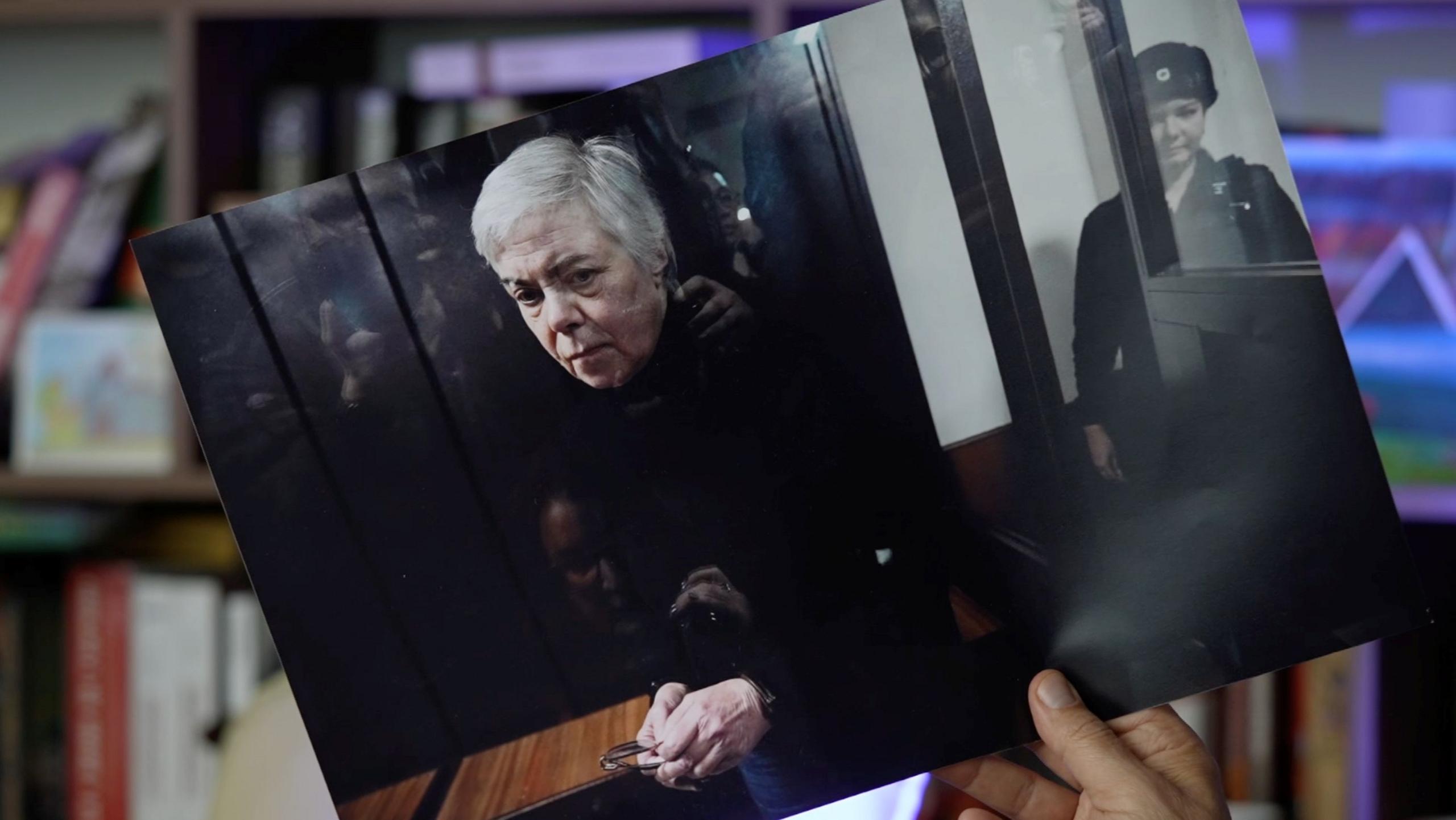 A hand holds a printed out photograph of Nadezhda Buyanova, a 68-year-old Ukrainian-born paediatrician in the dock at court, guarded.