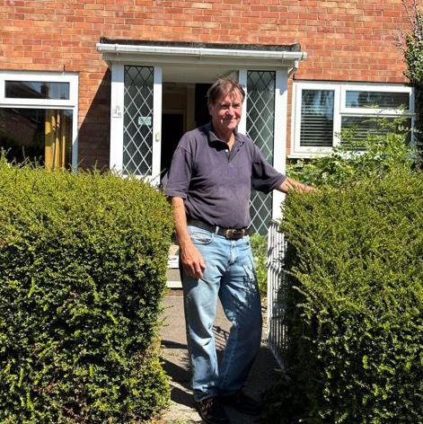 A man with brown haor stood next to hedges in front of a brick house.