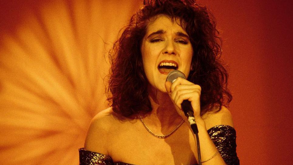 A young Celine Dion sings while holding a microphone. She is wearing a glittery strapless dress.