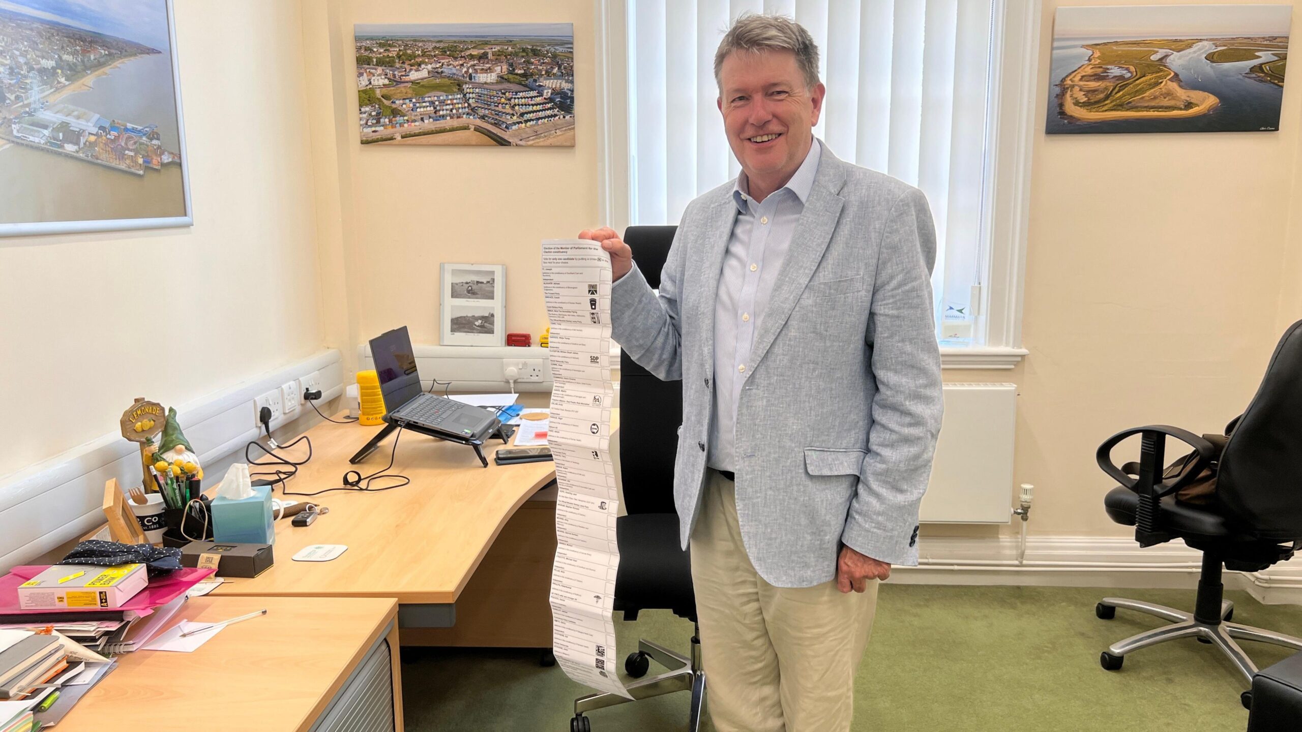 A man wearing a light blue linen jacket and light blue shirt, holding up a long white piece of paper that is thin and long like a giant receipt in an office.