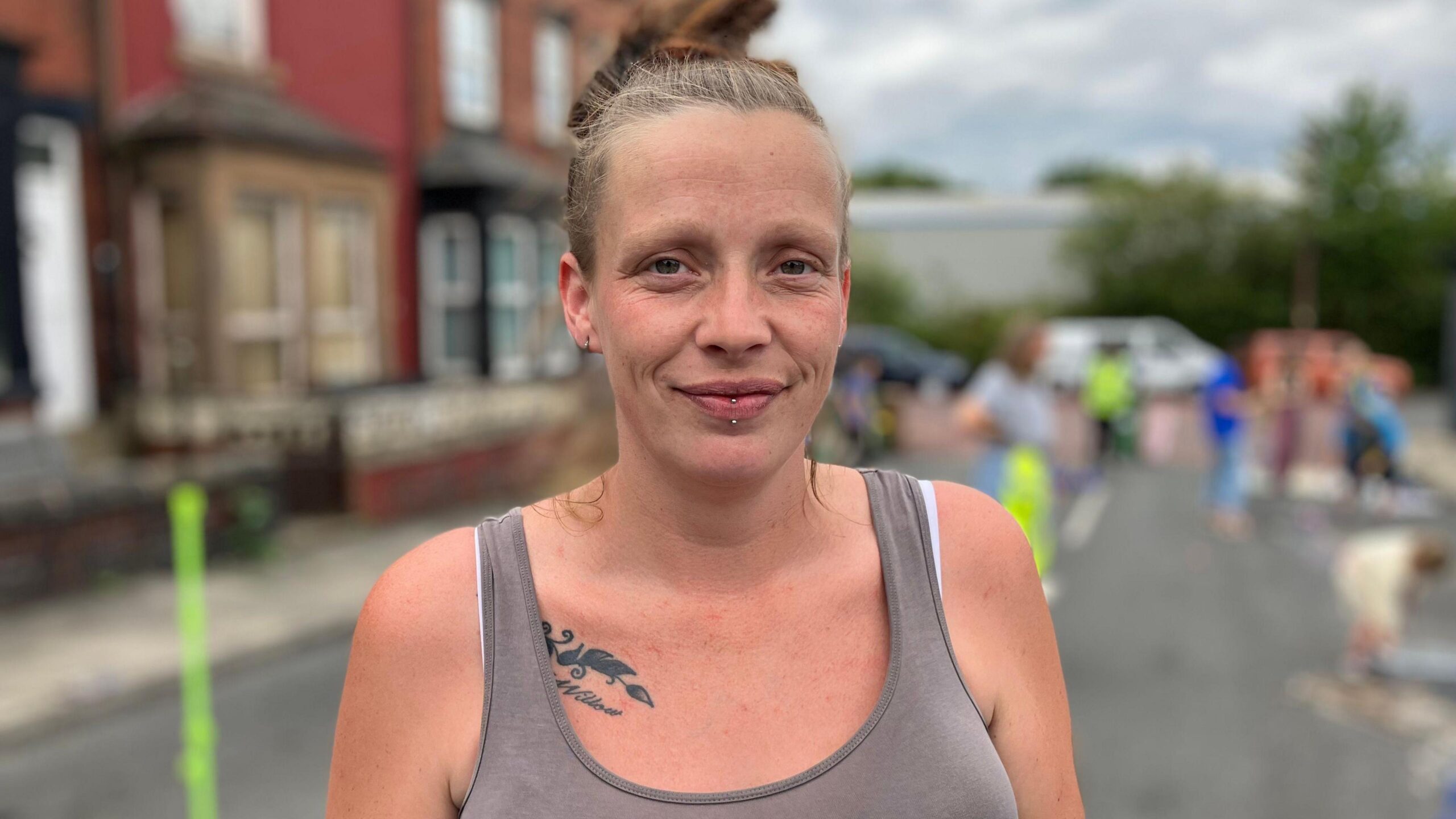 A white woman wearing a grey vest top is smiling at the camera. She is wearing her blonde hair up in a bun. Behind her children are playing in a residential street that has been blocked off to traffic.