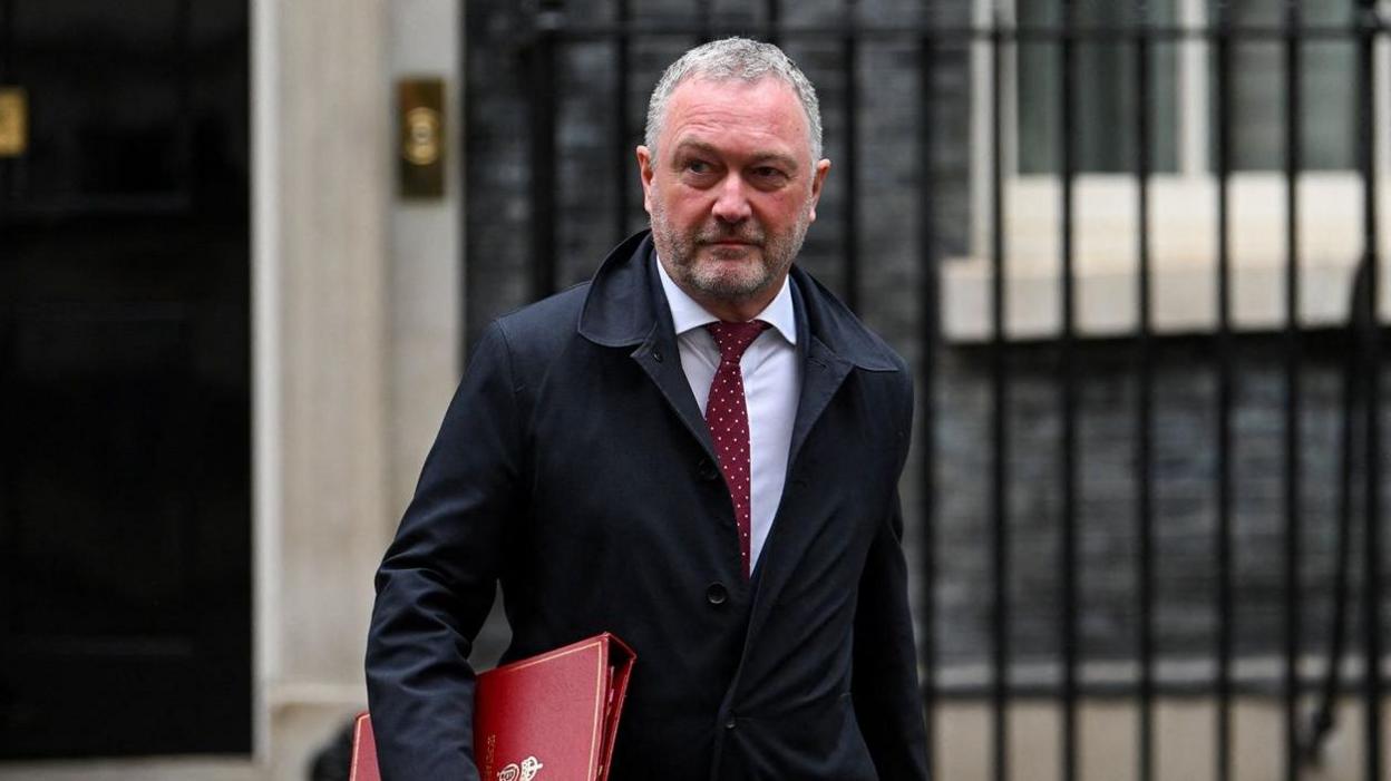 Steve Reed in a dark overcoat, white shirt and red tie walks outside a government building, carrying a large red document folder under one arm. The background features stone walls, a doorway and black metal railings. The photograph is taken at street level with the person in sharp focus and the building softly blurred behind.