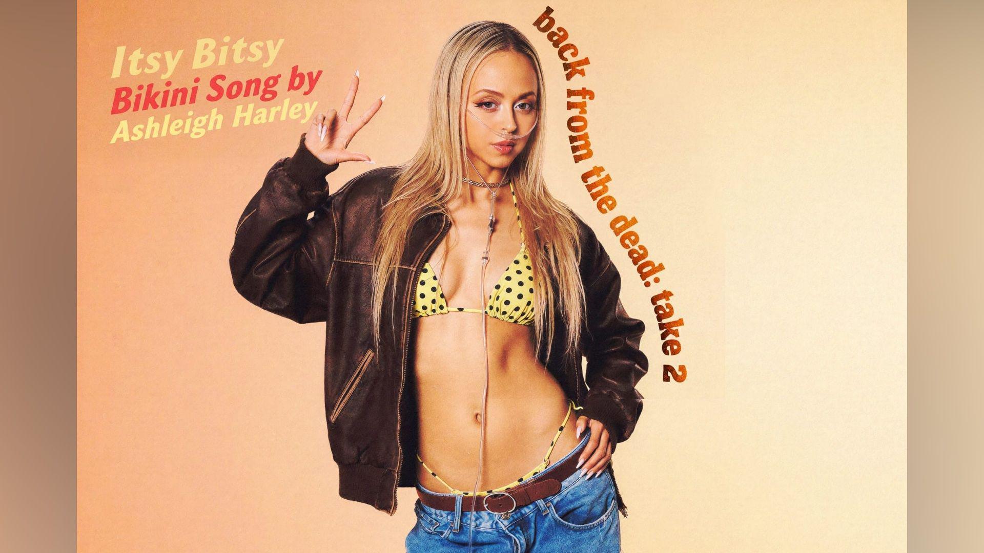 Harley, wearing a black leather jacket, a yellow bikini top and blue jeans, holds up a peace sign and places her other hand on her hip.
