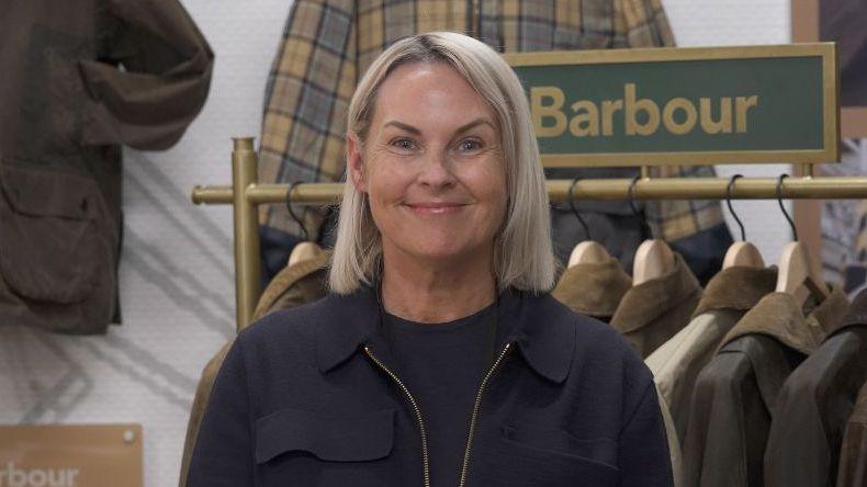 Woman standing in the foreground, wearing a dark jacket and top, facing the camera inside a clothing retail display. Behind the person is a rail of waxed jackets on wooden hangers and a green sign reading “Barbour”. Additional jackets hang on the wall, including a checked garment. A plaque on the left displays the text “Barbour Re-Loved”. The setting appears to be a branded clothing shop or exhibition area.