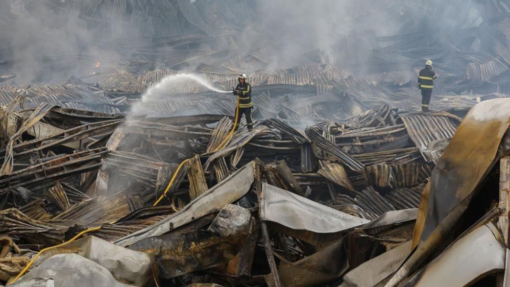 Rescuers work at the site of a Russian missile strike on a goods storage in Kyiv. A large stream of water can be seen coming from a hose one of the individuals is holding.