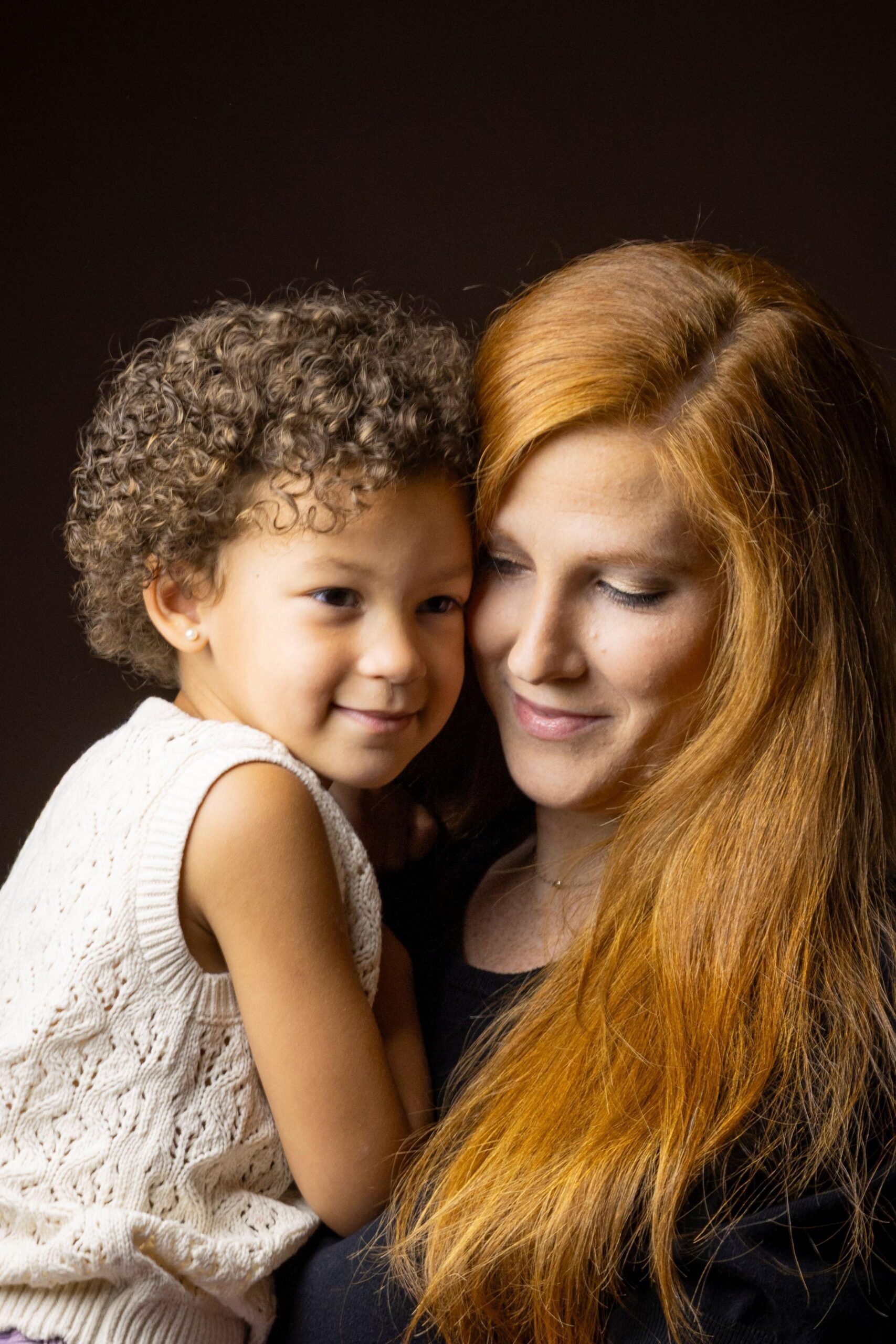 Close-up studio portrait of an adult with long red hair holding a young child with short curly hair. The child wears a sleeveless cream knit top, and both are positioned closely together against a dark brown background. Warm lighting highlights the hair texture and clothing details, creating an intimate family portrait.