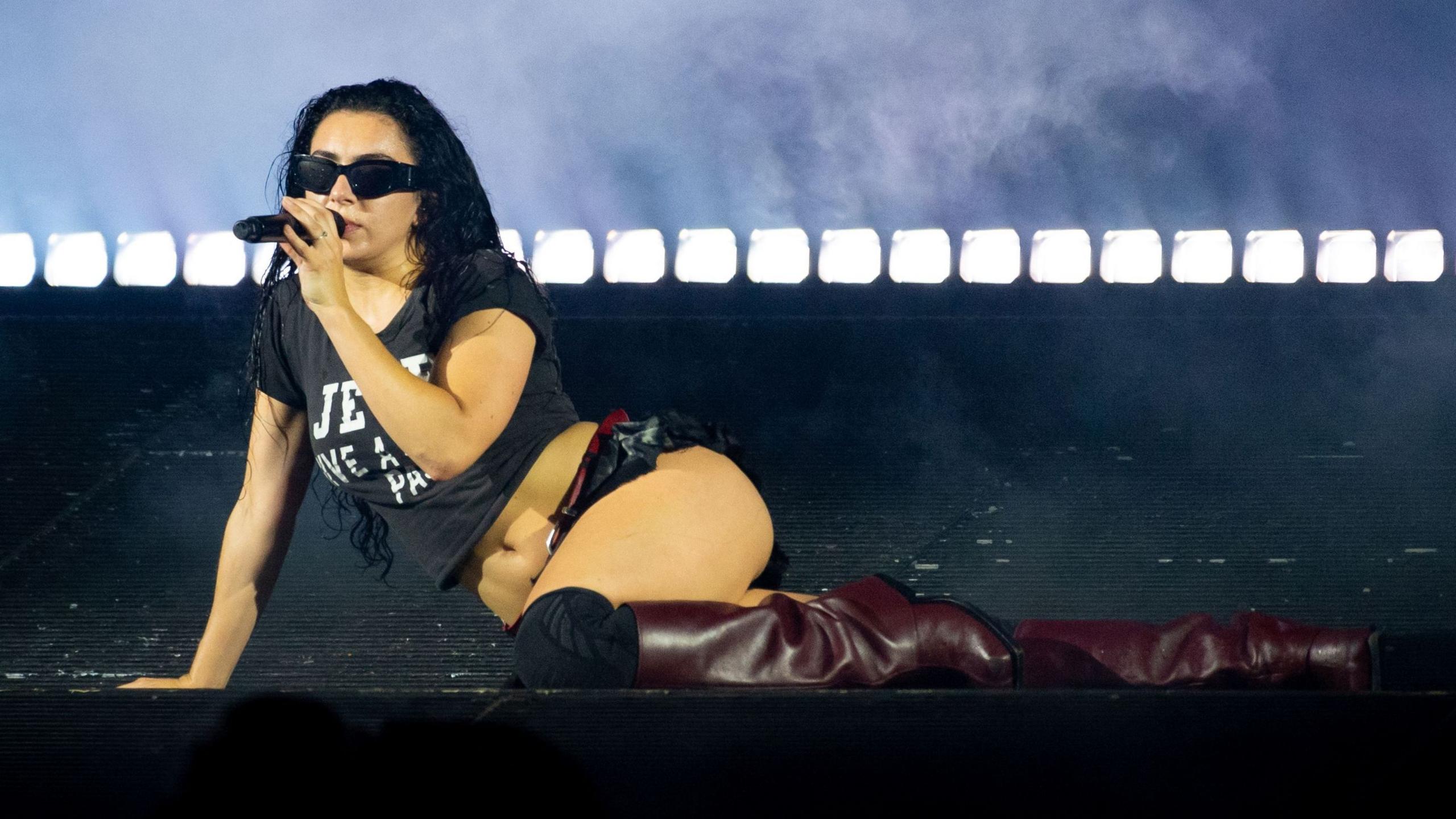 Charli XCX performing on stage. She is holding a microphone and wearing a black t-shirt, a black short skirt and red boots.