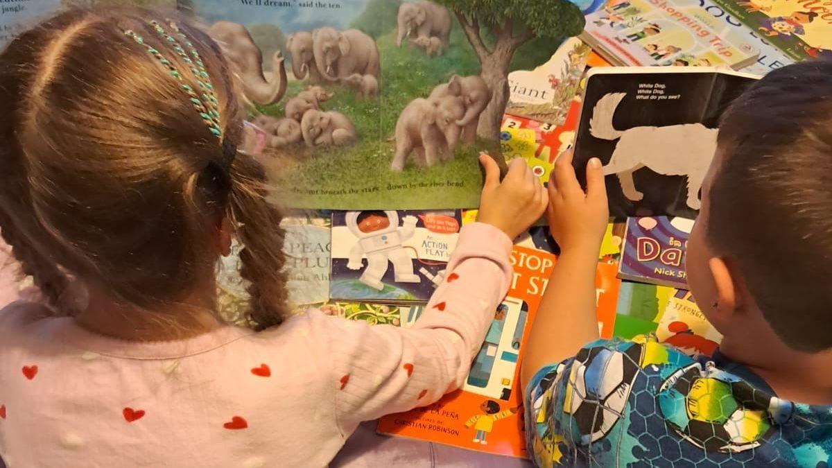 Two children in pyjamas, their backs turned to the camera, each hold books open - atop a pile of other books they received from Parton's Imagination Library