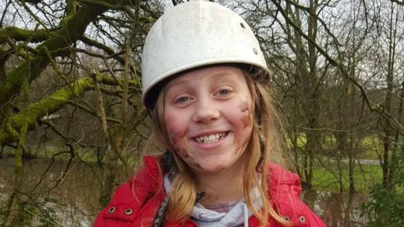 A younger Menna smiles at the camera. She has long blonde hair, wears a white helmet and red coat, and has mud on her cheeks. A river and trees can be seen in the background.