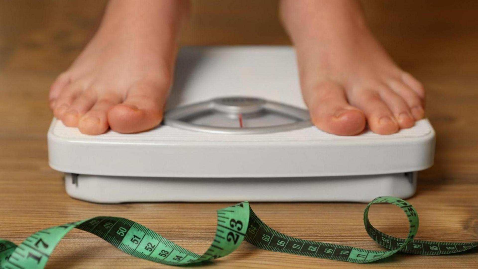 Person on scales in bare feet. The scales are white with a red needle and there is a green fabric tape measure on the wooden floor in front of them.