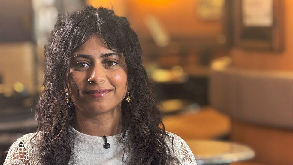 A portrait shot of Taiyaba Zeria who has long dark brown hair and is wearing a white top. She is staring at the camera in the cafe of the