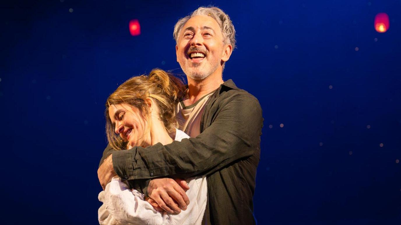 Two performers stand together on a dimly lit stage, framed by a deep blue backdrop and glowing red lanterns suspended overhead. Warm spotlighting highlights the embrace, while the floating lights create a dreamlike, theatrical atmosphere.