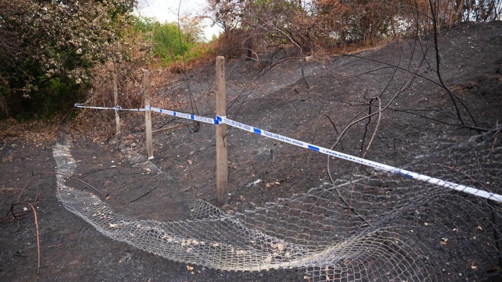 Area of darkened ground and sparse vegetation cordoned off with blue-and-white tape and temporary fencing on a sloping embankment.