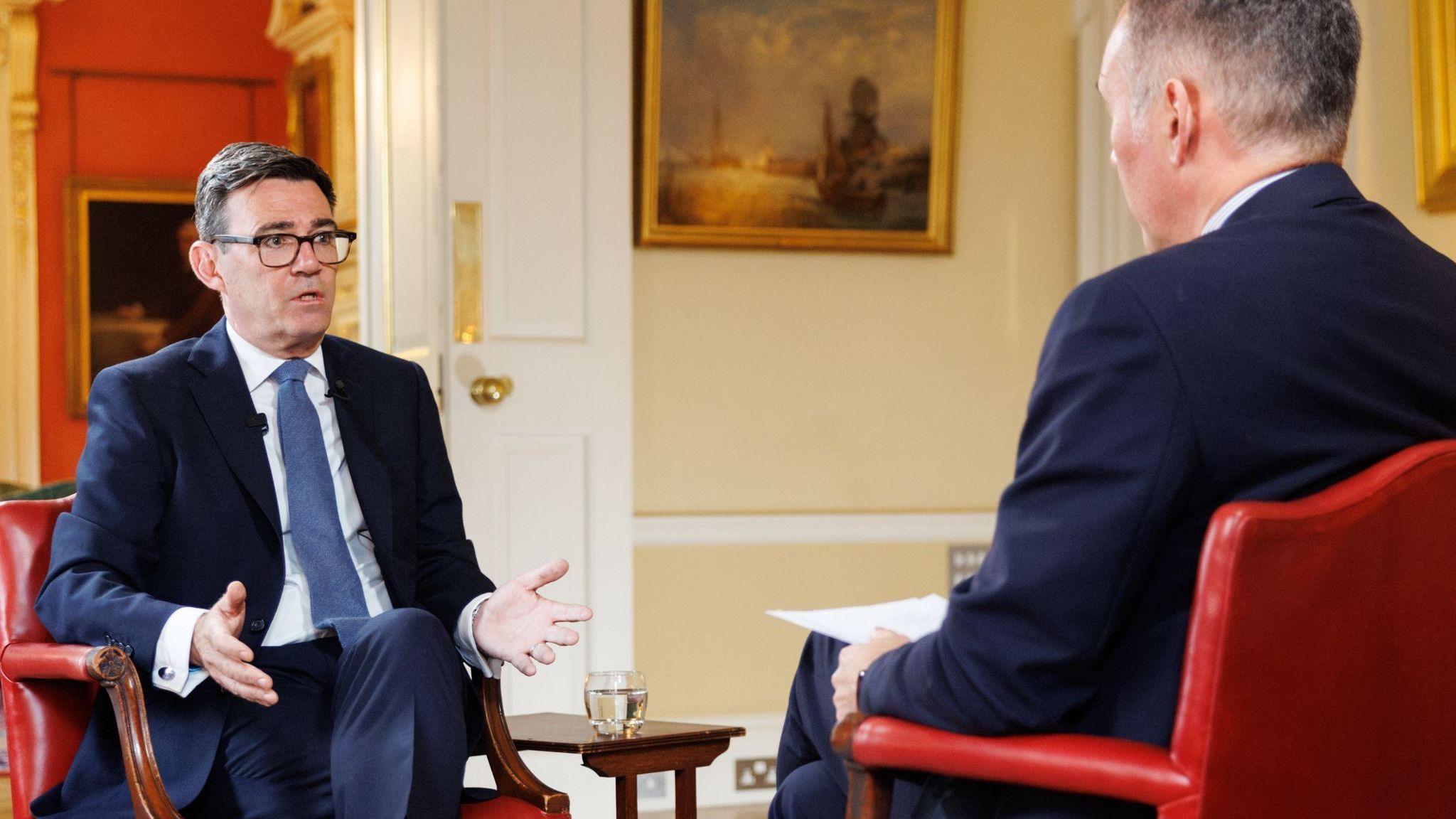 Andy Burnham wears a dark suit sitting on a chair speaking with Roger Johnson who wears a dark suit. They are both sitting on red chairs.