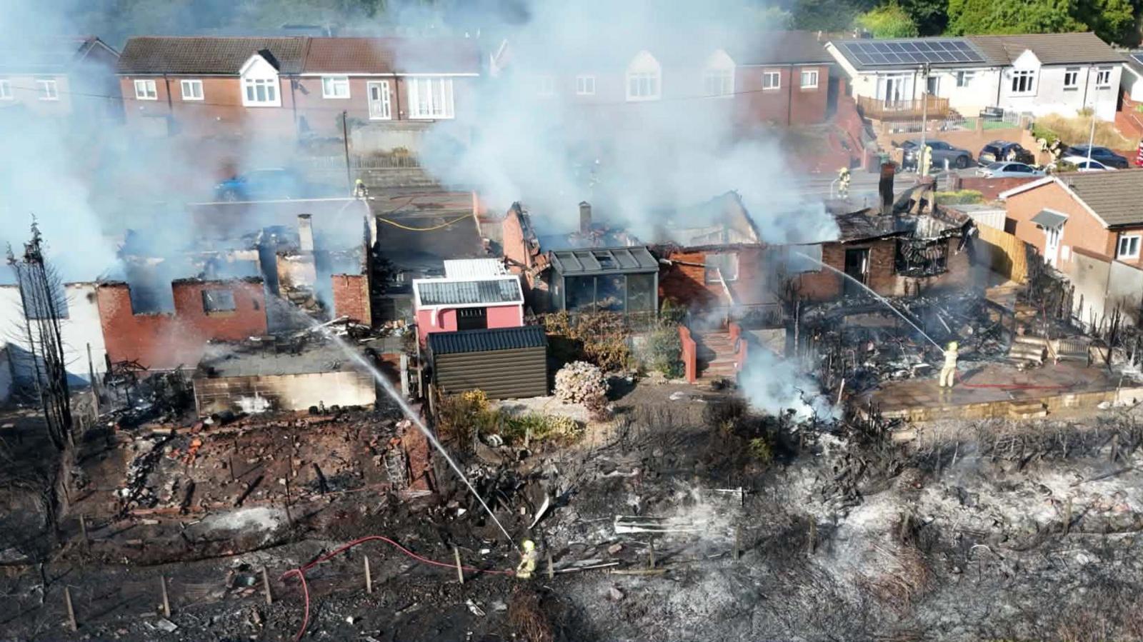 An aerial shot of a row of homes with their roofs destroyed by fire. Smoke streams into the air. Land in front of the houses is scorched, with firefighters holding hoses spraying water towards the scene.