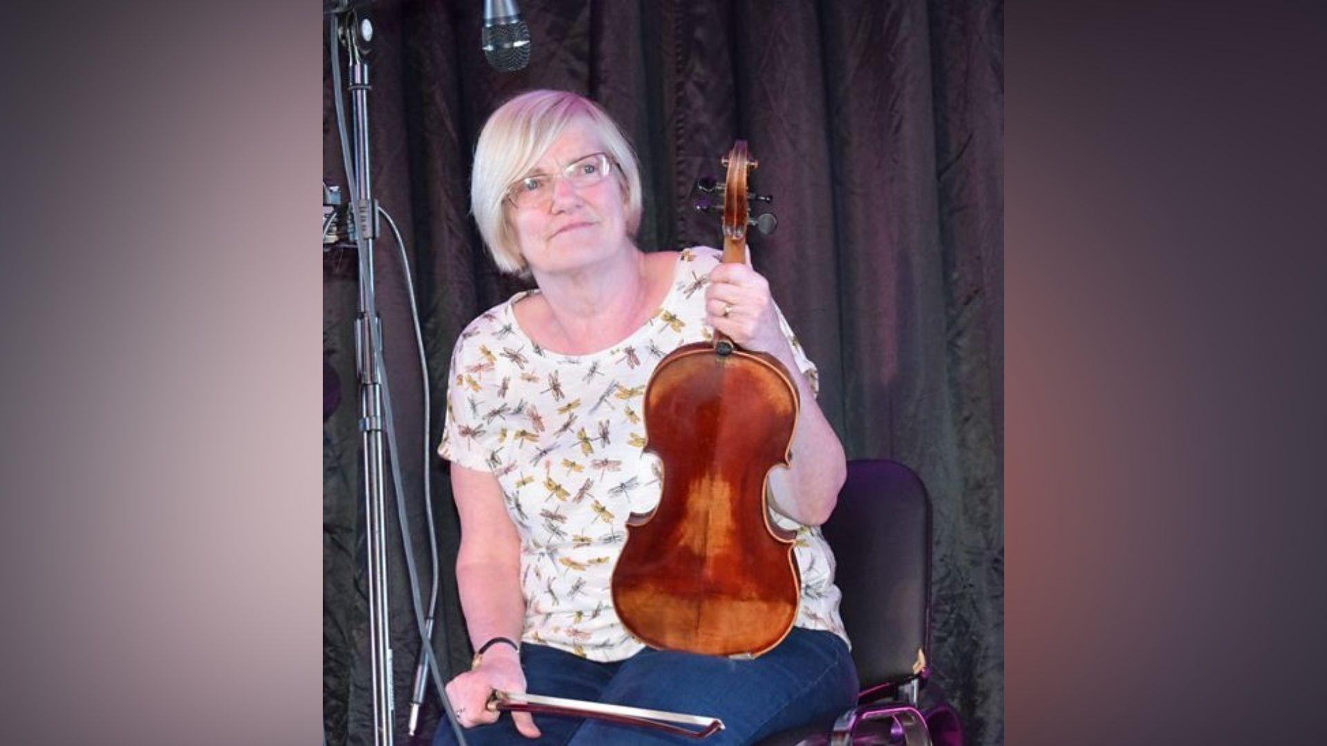 A woman with short white hair is sitting on a black chair. She is holding a violin and a bow. She is wearing a white t-shirt with butterfly's on it.