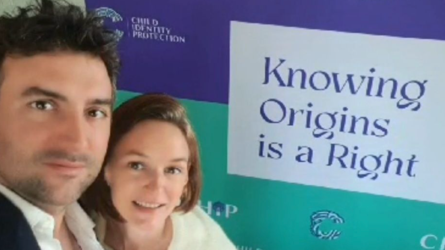 Audrey and Arthur stand close together in front of a large campaign banner which displays the message “Knowing Origins is a Right” and includes logos associated with child identity advocacy.