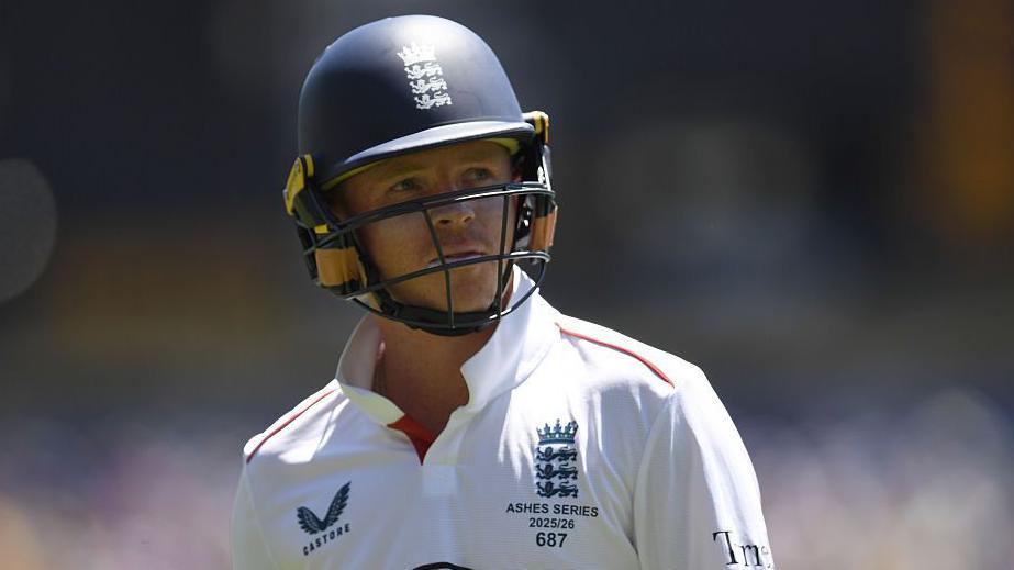 Ollie Pope, wearing a blue helmet and white England shirt, looks to his left