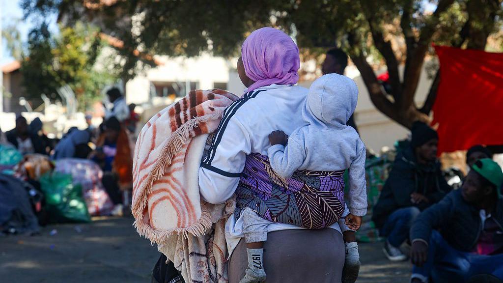A woman with a baby strapped to her back is facing away from the camera. She is holding a warm blanket and wearing a mauve headscarf. The baby is wearing a grey hoodie. Some others can be seen out of focus in the background.