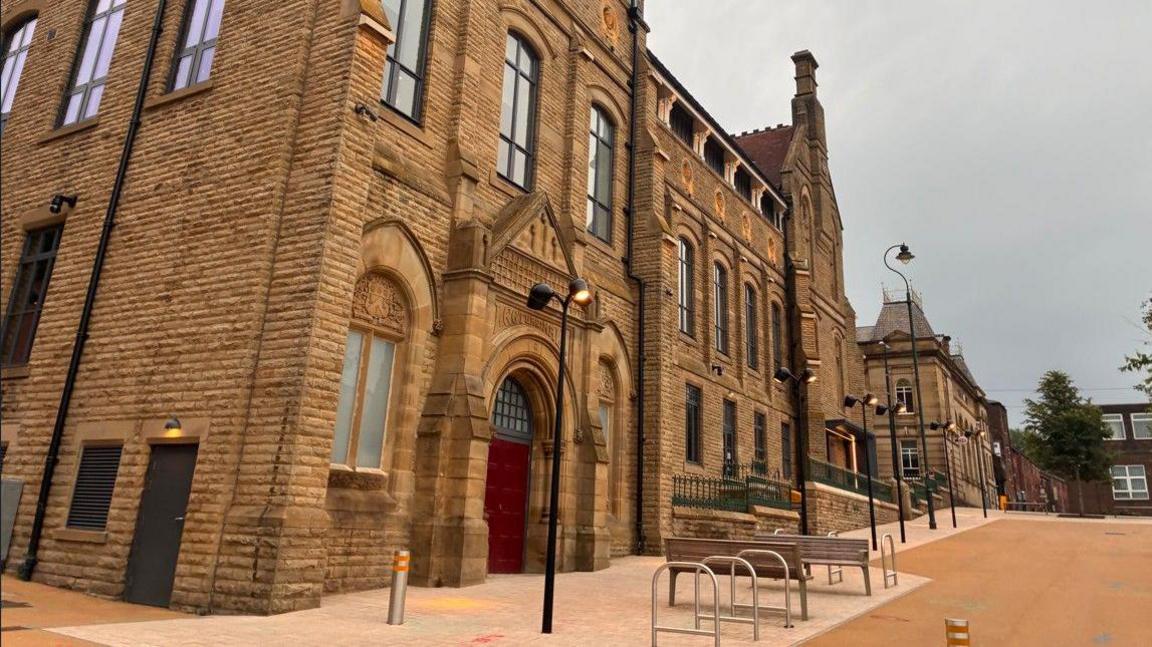 A historical pale brick building with a red door towers above a public square on an overcast day.