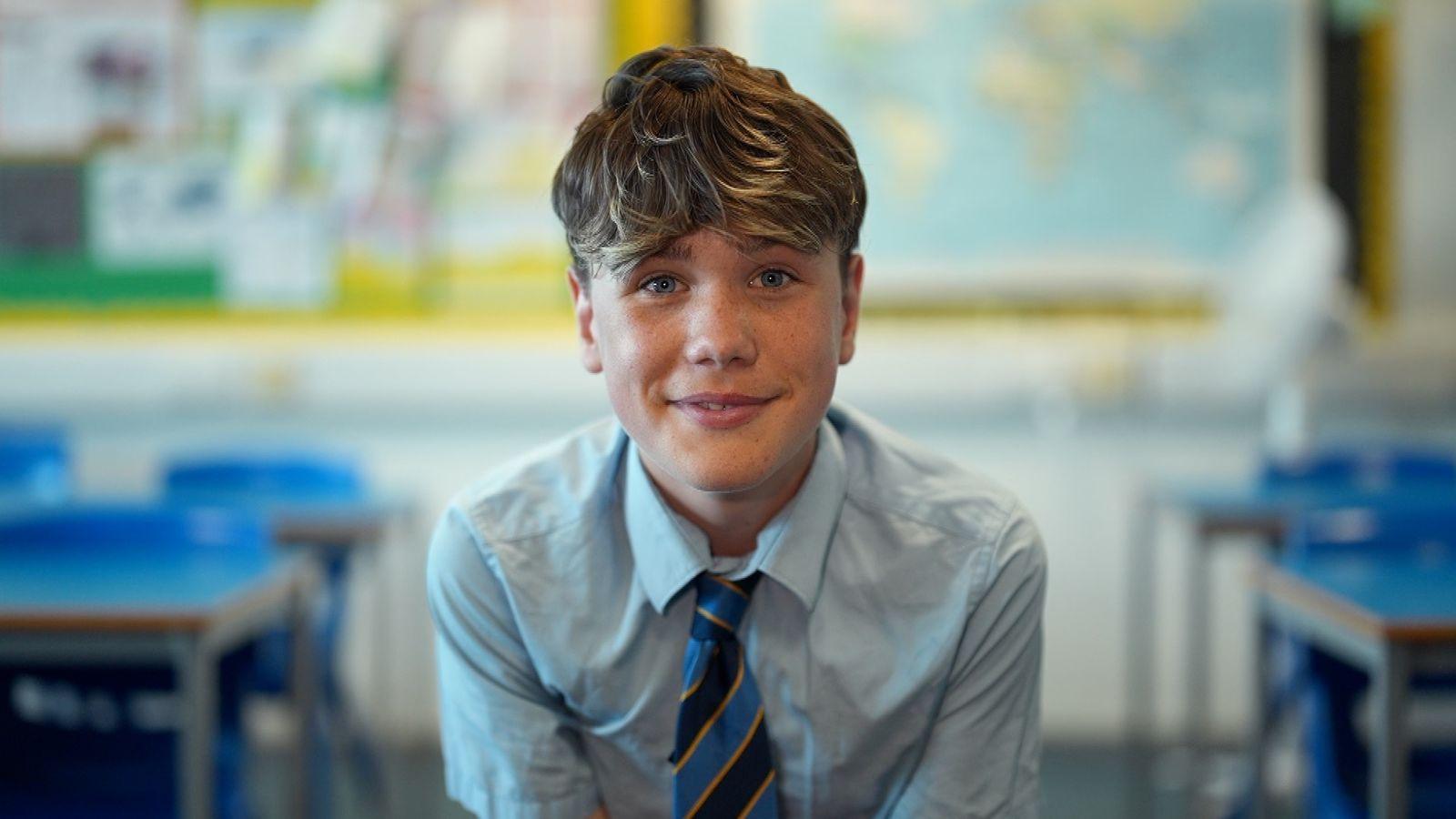 A young school pupil, Ethan, smiles as the camera. He is wearing a school uniform - black, yellow and blue striped tie and pale blue shirt - and sitting in a classroom.
