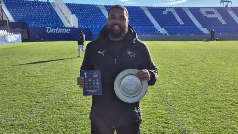 Derby County Under-12s lead coach Zeph Thomas with the trophy after his side  won the Orange Veins Madrid tournament in 2025