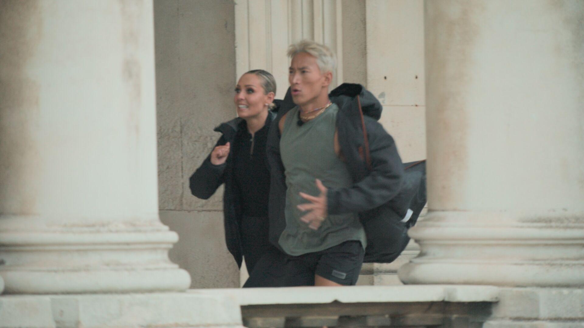 Carlos Gu and Amy Dowden pictured running side by side during an episode of the Channel 4 show, Celebrity Hunted. Carlos is wearing a grey dark coat, a light grey t-shirt and several necklaces. Amy is wearing a black top, jacket and coat. They are both not looking at the camera.