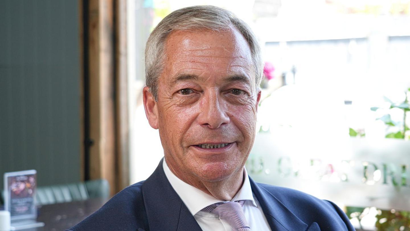 Nigel Farage sitting in a pub while wearing a white shirt and navy blazer. He is smiling.