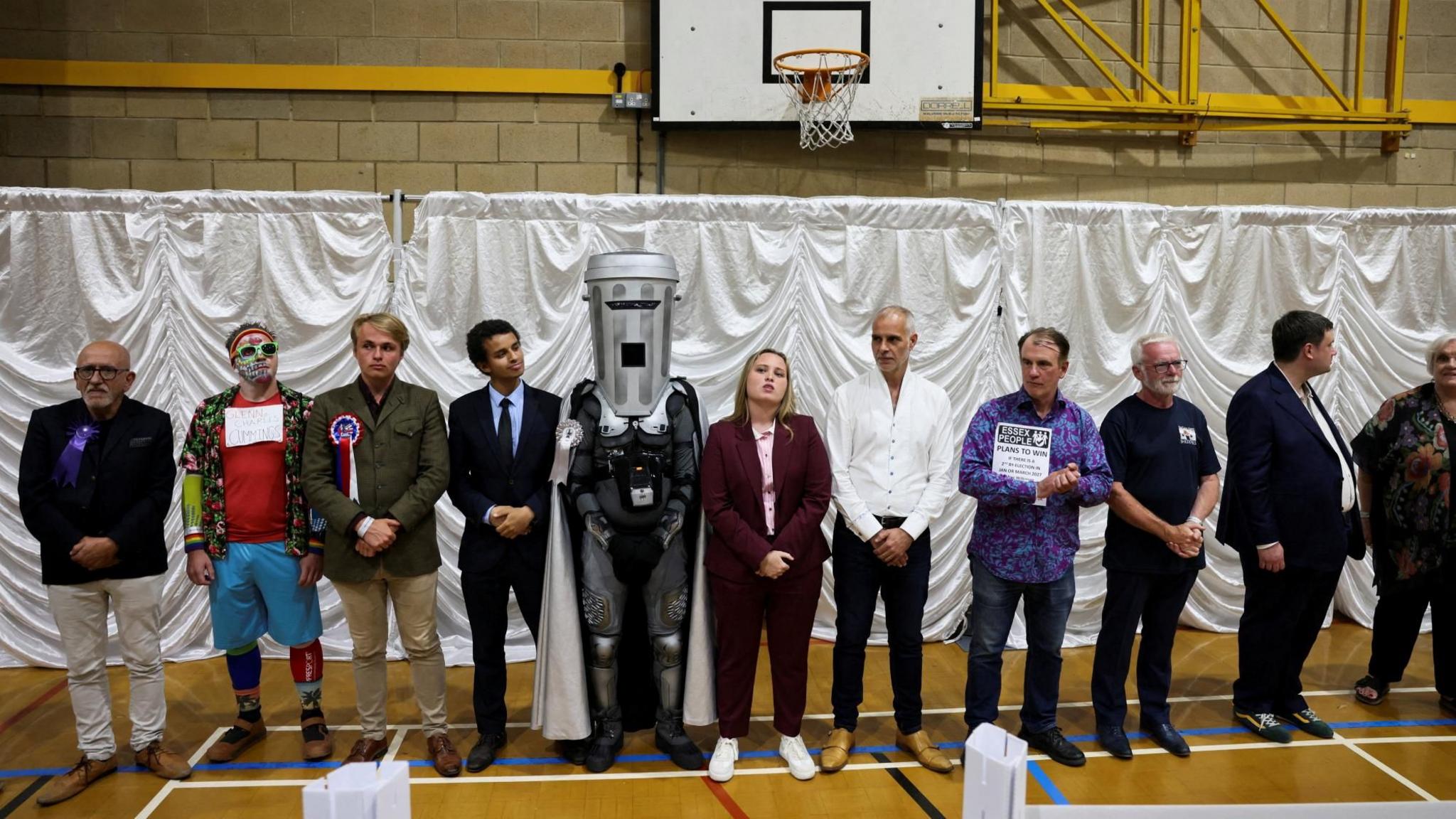 Eleven of the candidates at the 2026 Clacton by-election stand in a line on the floor of a sports hall. Count Binface is prominent in the middle, wearing his dustbin headgear.