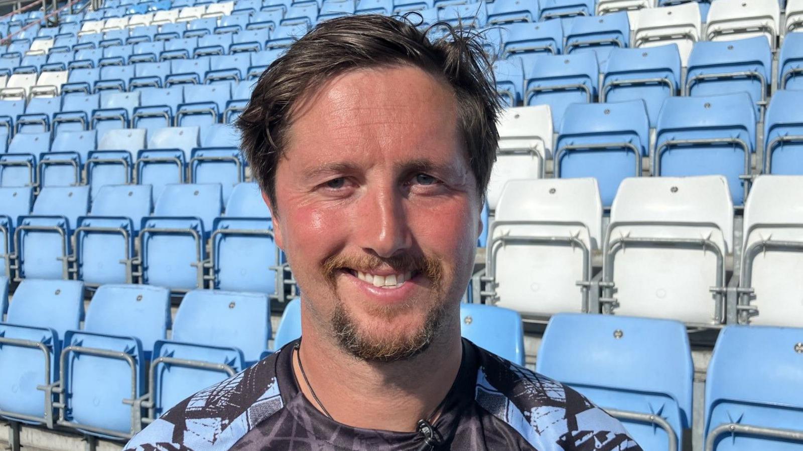 Oli Bishop smiling in front of an Exeter Chiefs stand of seats, wearing sky blue and black training top