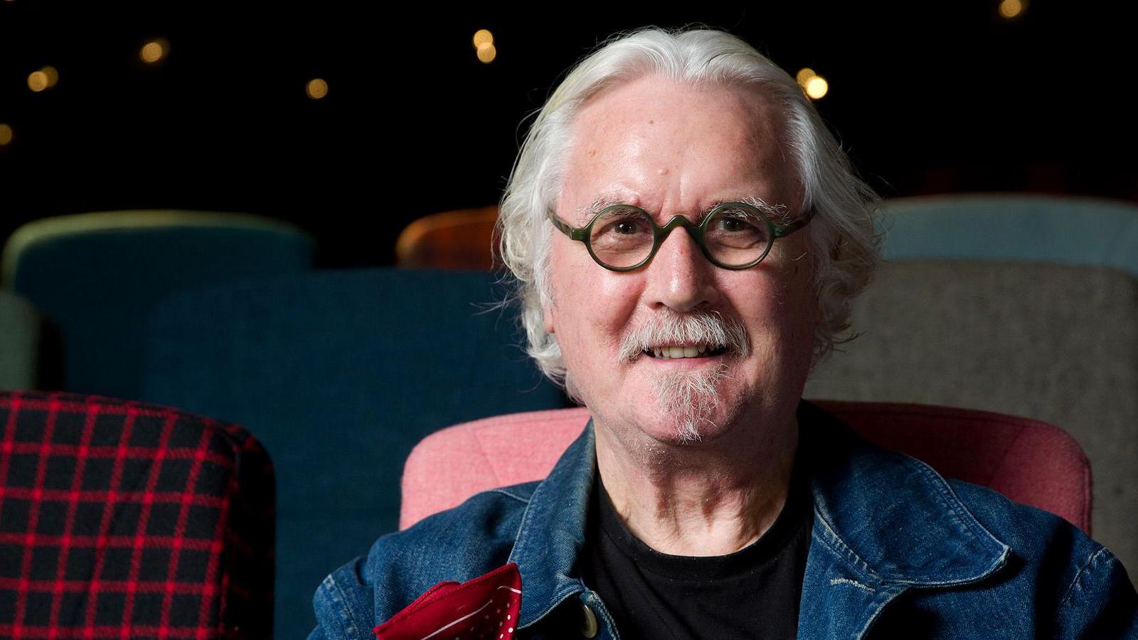 Sir Billy Connolly sitting in a theatre with colourful upholstered chairs in the background. He is wearing a denim jacket with a patterned red cloth in the pocket.