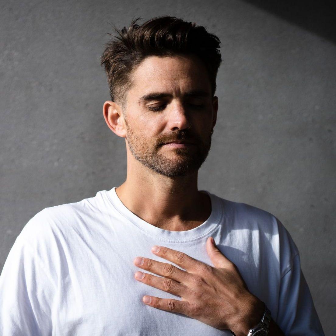 Person wearing a plain white T-shirt standing in the foreground with one hand placed on their chest. Their eyes are closed and a wristwatch is visible on one wrist. The background consists of a plain grey wall, with directional light creating areas of shadow across the wall and part of the upper body.