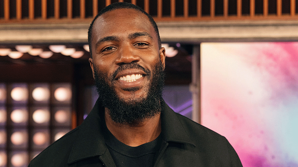 Mo Gilligan in black shirt and jacket smiles for the camera in a TV studio