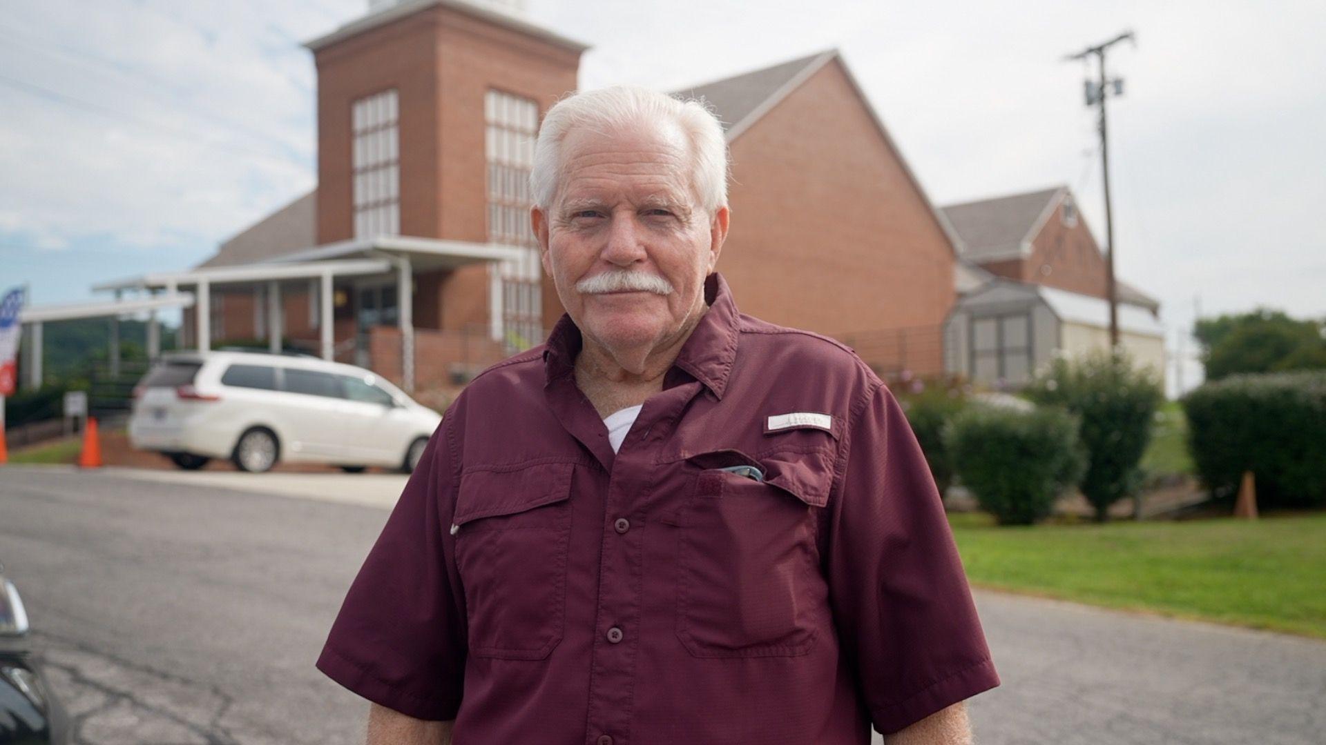 Seventy-five-year-old Willam Bogle, sporting white hear and a full white mustache, wears a maroon button-up shirt as he stands in front of a brick church