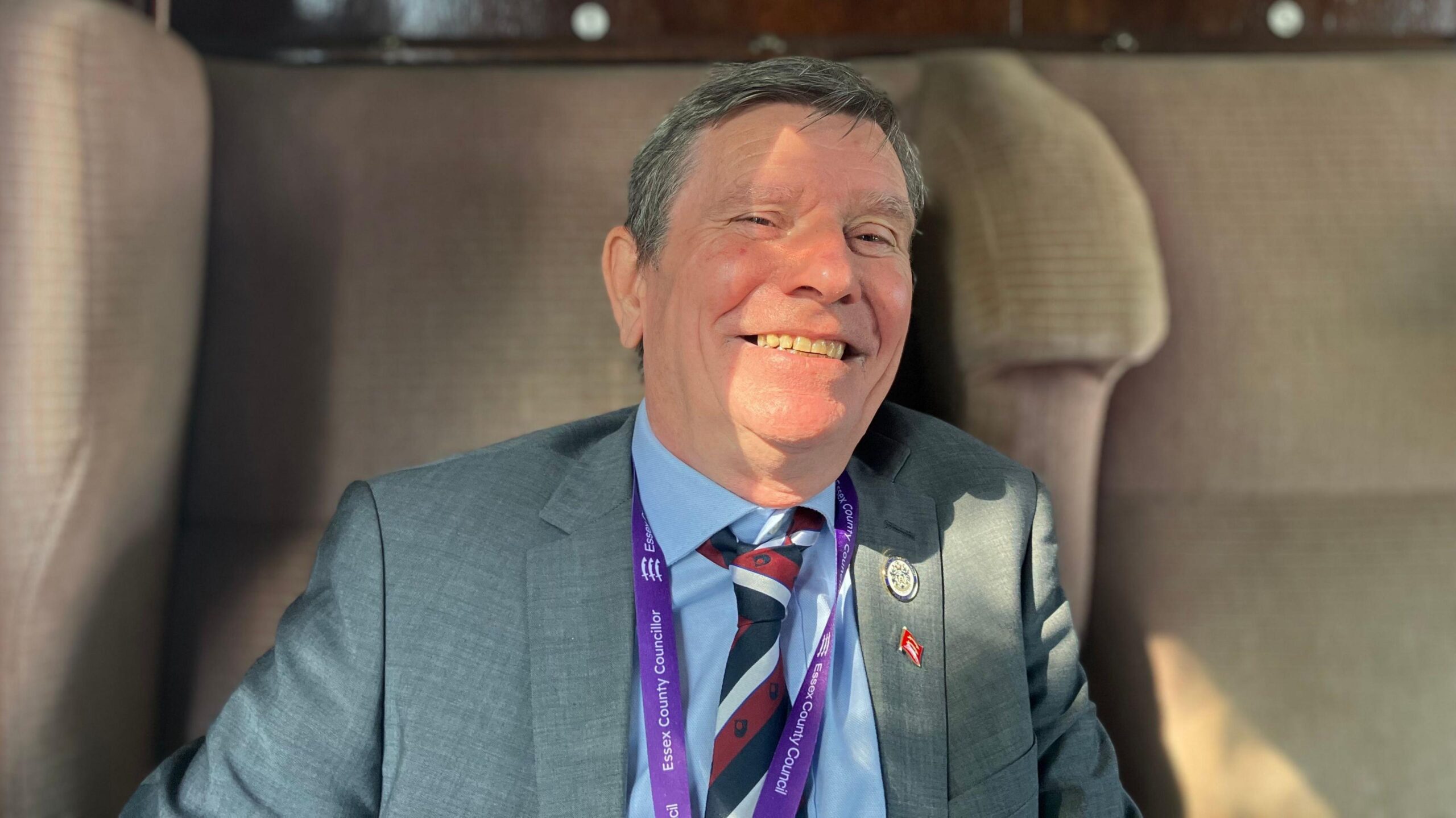 Jeff Bray has greying dark hair and is wearing a grey suit, blue shirt and striped tie. He is smiling widely at the camera and is sitting in a beige train seat.