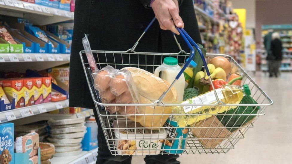 Person holding a wire shopping basket in a supermarket aisle containing bread, eggs, milk, fruit, vegetables, and packaged groceries, with store shelves visible in the background.