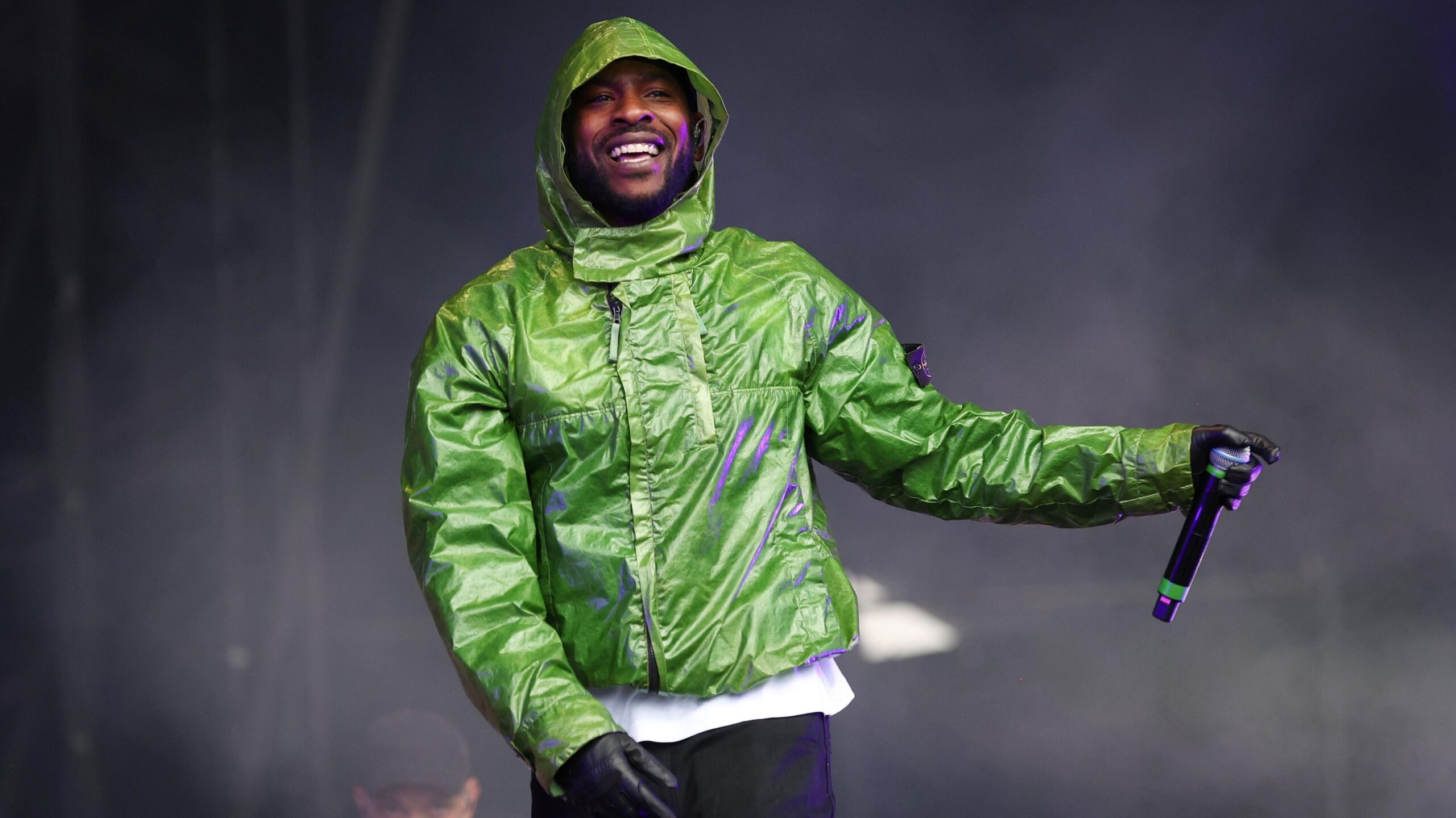 Skepta performs on stage wearing a green jacket, black gloves and black trousers.