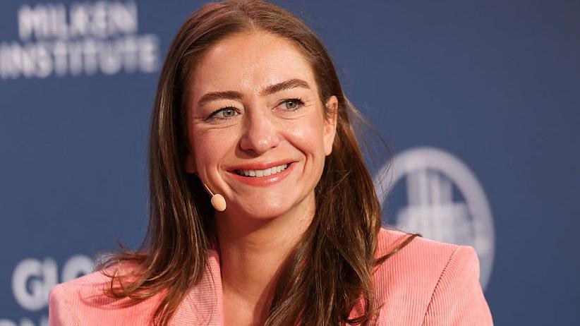 Whitney Wolfe Herd smiling while wearing a pink corduroy blazer at the Milken Institute Global Conference against a blue backdrop.