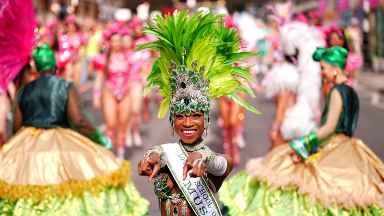 A woman in a green beaded costume and feathered headdress points at the camera with a smile.