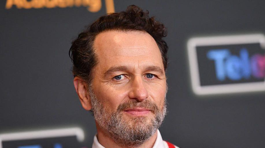 Rhys at a press junket wearing a white sweater with red and blue stripes and a white shirt with a red tie