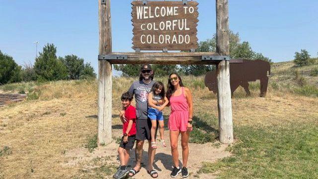 Colorado mother Ciara Fernandez Faber, wearing a short pink romper, stands with her husband, son and daughter under a sign that reads 'Welcome to Colourful Colorado' - with grass, shrubbery and a bison art piece in the background